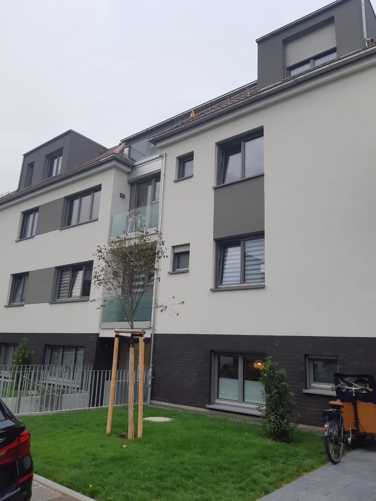 Modern apartment building with white and black exterior, mansard roofs, and green lawn with young tree in front